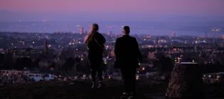 Two people facing away from camera overlooking an Edinburgh skyline at twighlight. The sky is pink and purple