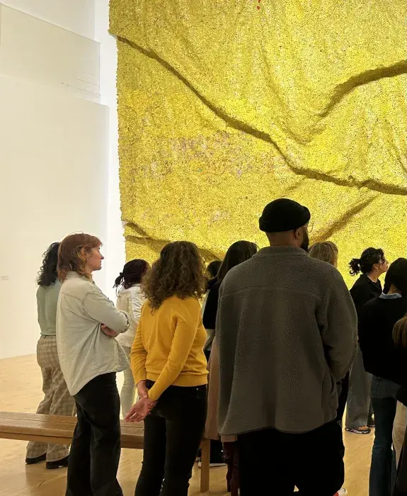 Participants of a Decolonised Transformations workshop afcing away from the camera looking at a large-scale piece of artwork which is golden yellow in colour.