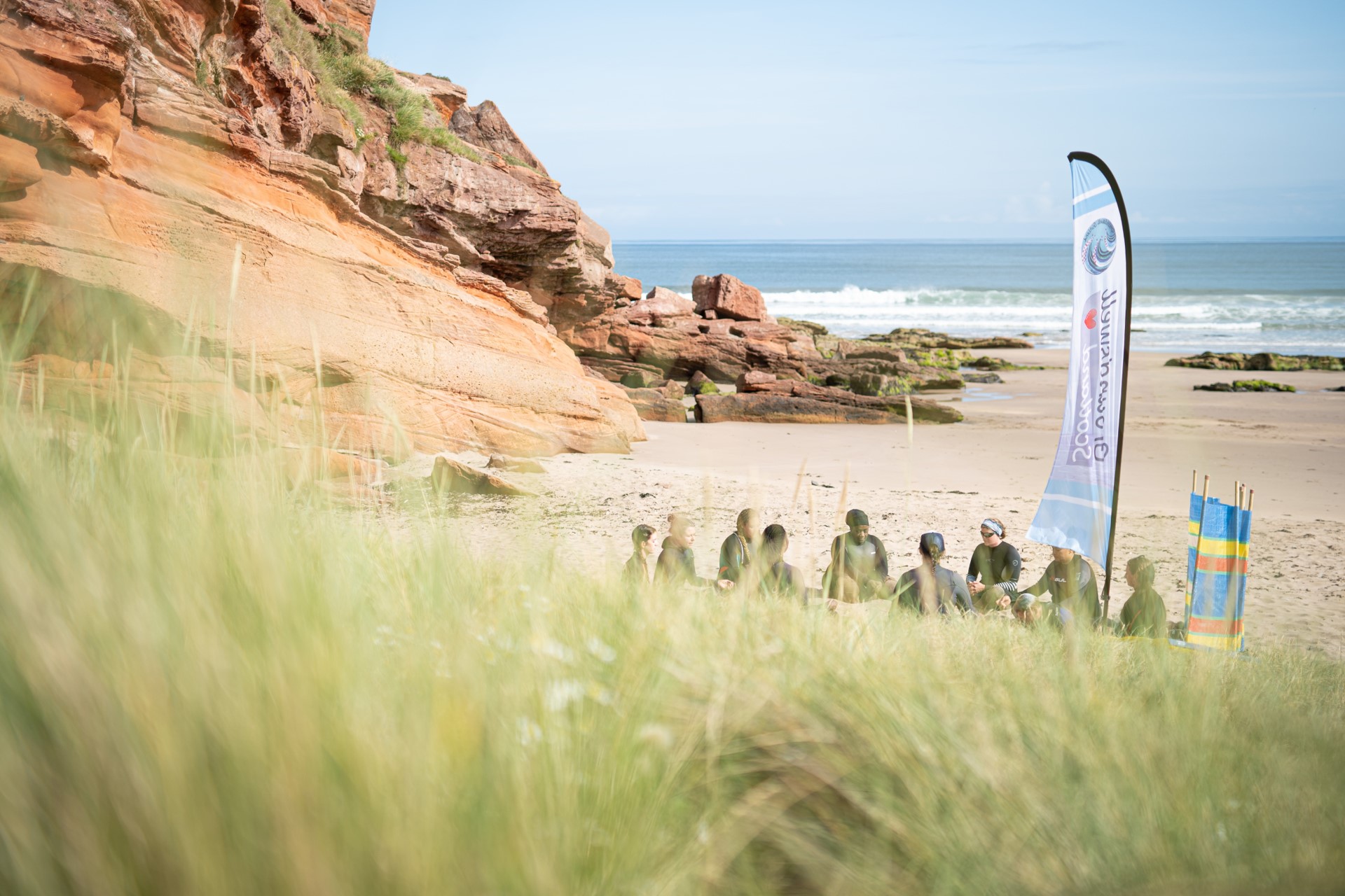 James Appleton: Groundswell Scotland: Women sitting on a beach in wetsuits with a Groundswell Scotland flag